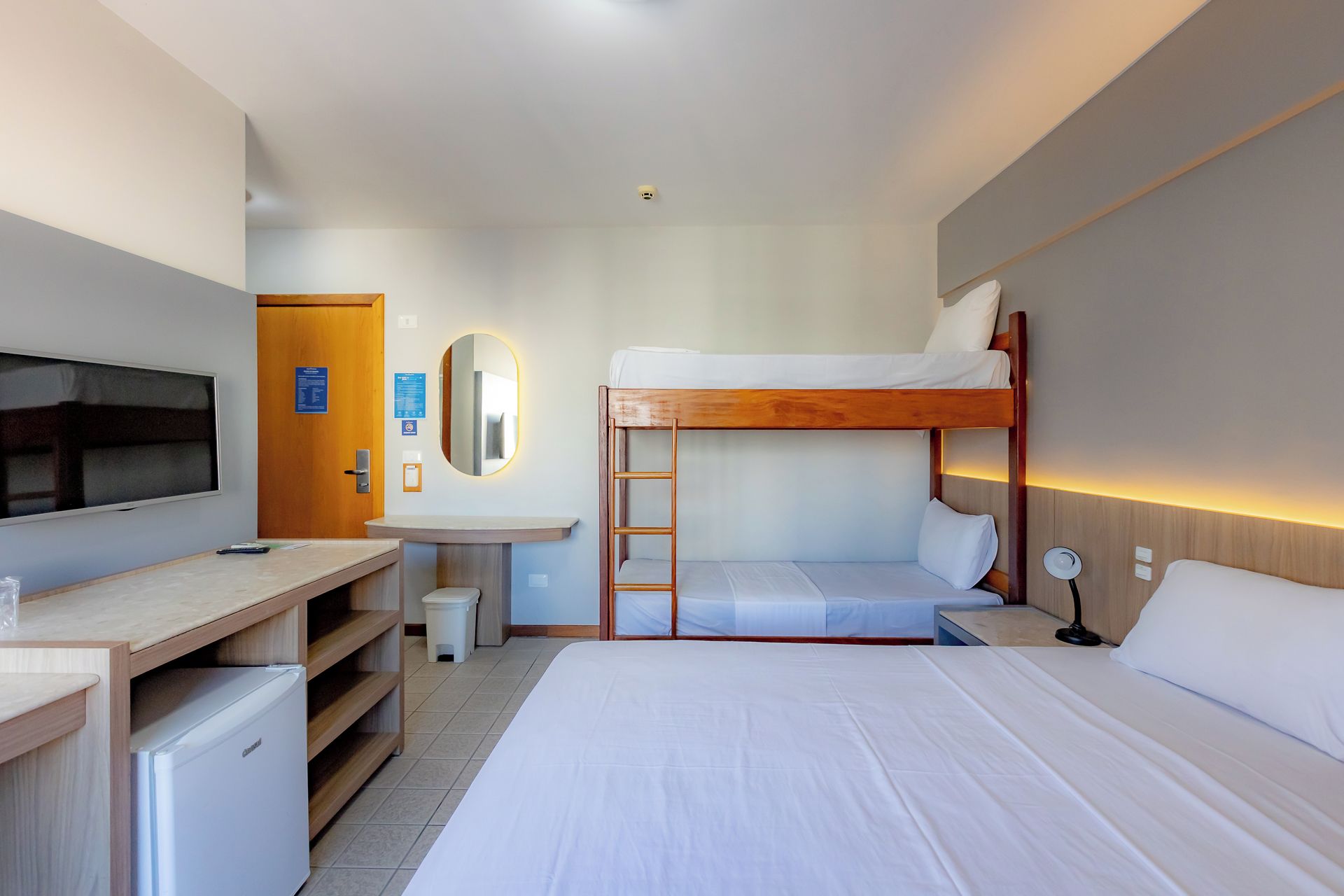 A hotel room with a bunk bed, a desk with a television, a mirror, and a bedside lamp.