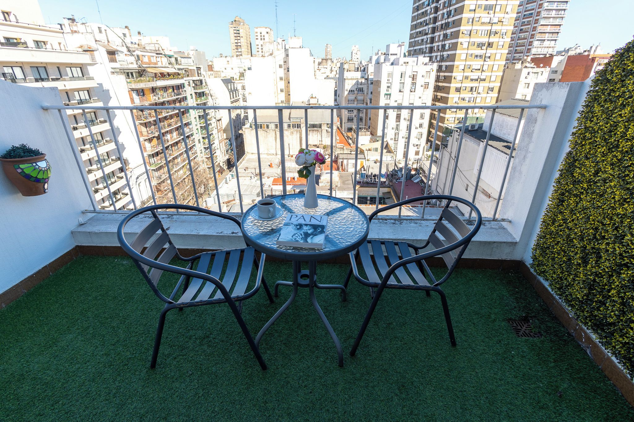 Un balcón con una mesa redonda y dos sillas, una planta en maceta y vistas de la ciudad.
