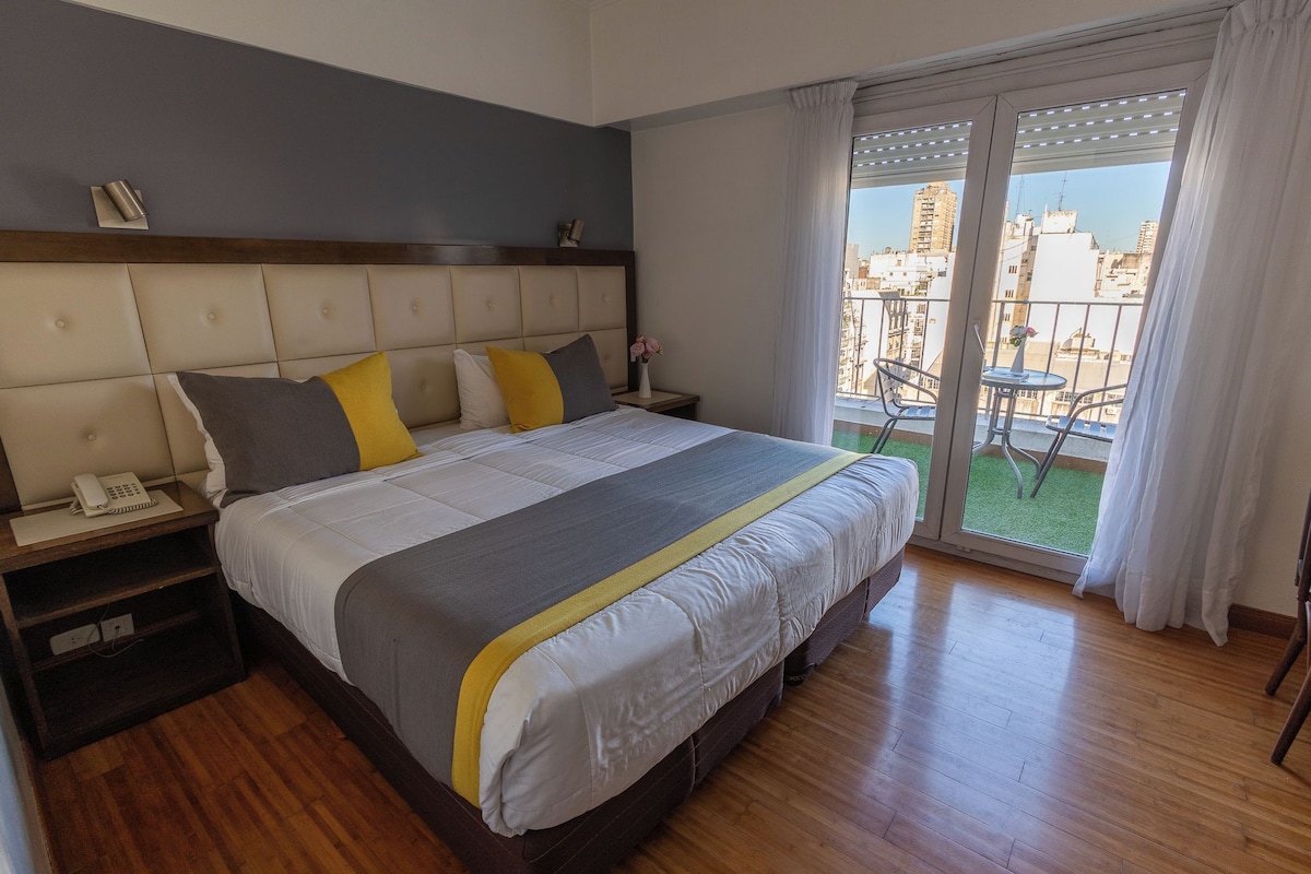 A hotel room with a large bed, a bedside telephone, a small table with a chair, and a view of the city through a balcony.
