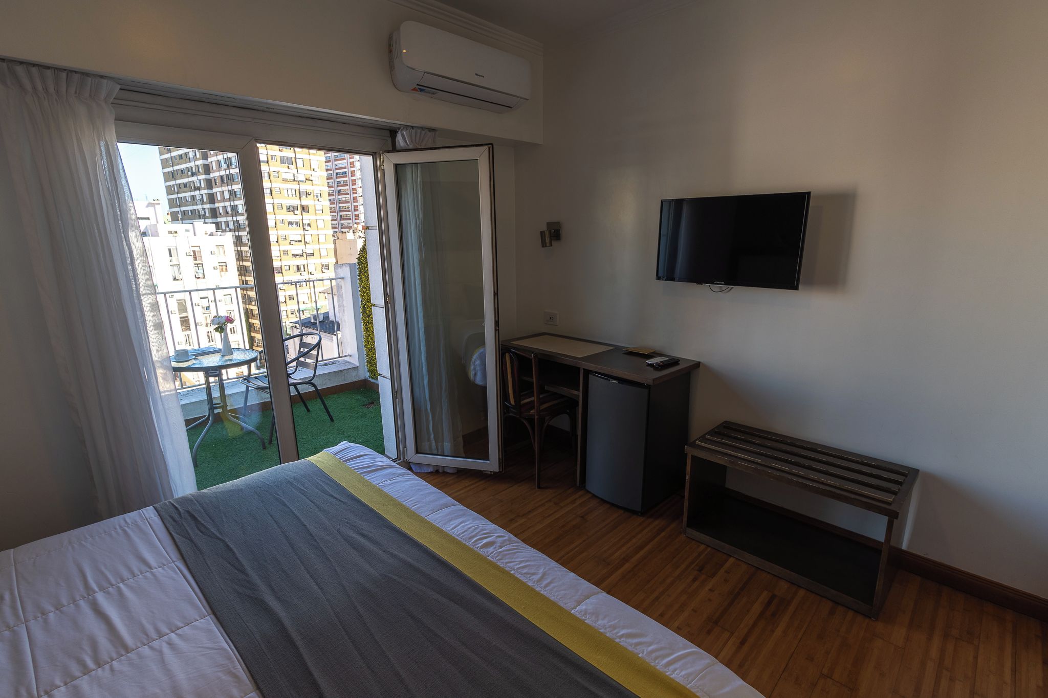 A hotel room with a bed, a desk, a TV, and a balcony with a view of buildings.