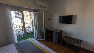 A hotel room with a bed, a desk, a TV, and a balcony with a view of buildings.