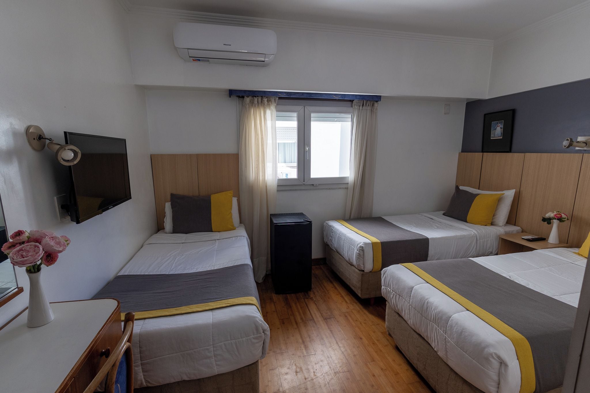 A hotel room with three beds, a TV, a window with curtains, and an air conditioning unit.