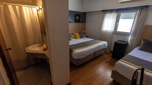 Quarto com cama, mesa de cabeceira, janela com cortinas e banheiro com cortina de chuveiro.