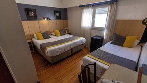Quarto de hotel com duas camas, uma mesinha, uma cadeira, janela com cortinas e luminária na parede.