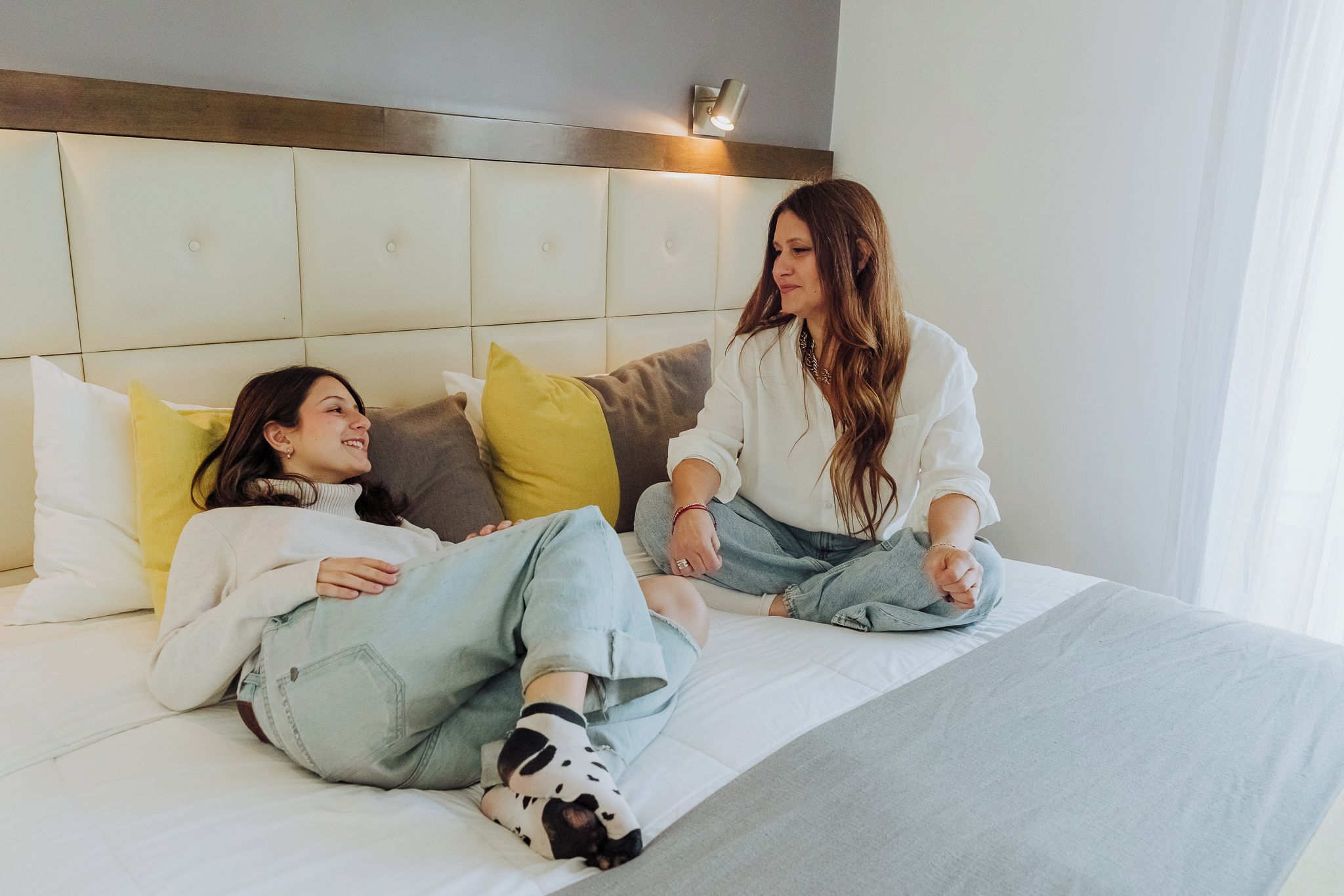Two people sitting on a bed with a headboard, pillows, and a light fixture above.