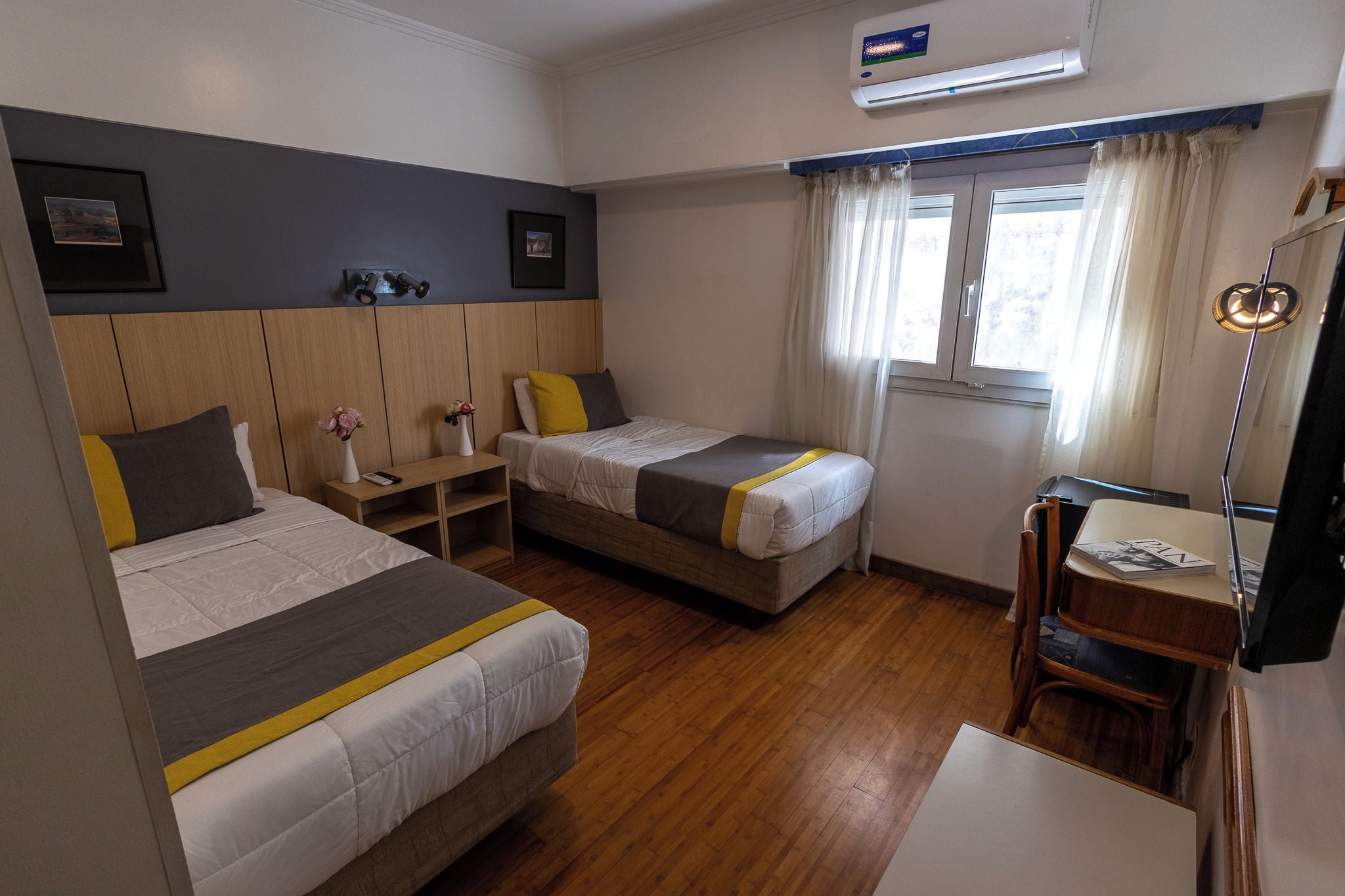 A hotel room with two beds, a desk, a chair, a lamp, and a window with curtains.