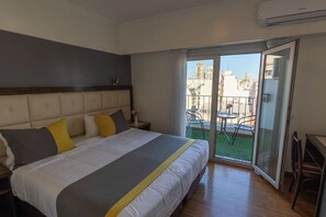 Quarto de hotel com cama, mesa de cabeceira, janela com vista para edifícios e varanda com cadeiras.