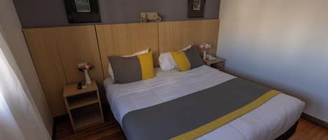 Una habitación de hotel con cama, mesitas de noche, lámpara de pared y cuadros enmarcados.