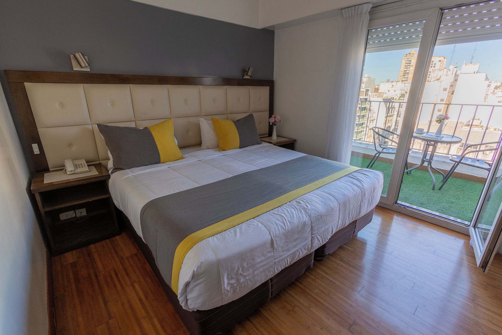 A hotel room with a bed, bedside tables, a phone, and a view of the city through a balcony.
