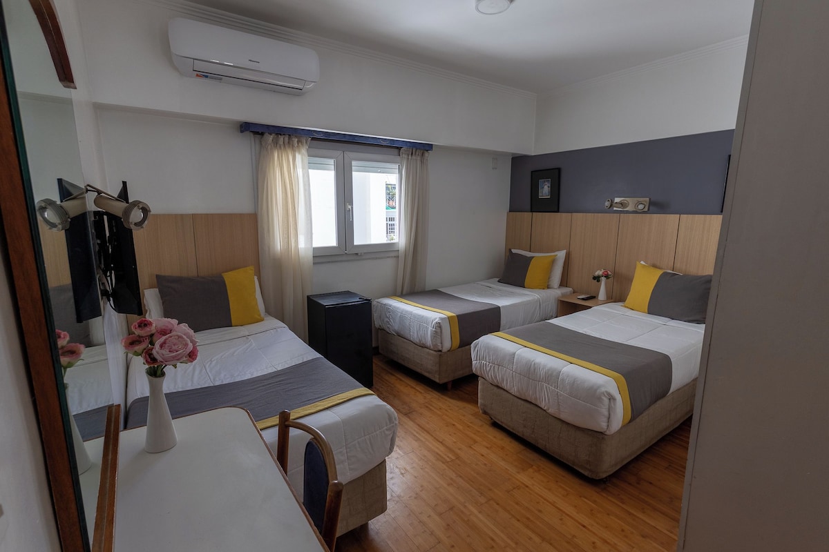 A hotel room with two single beds, a bedside table with a lamp, a mirror, and a window with curtains.