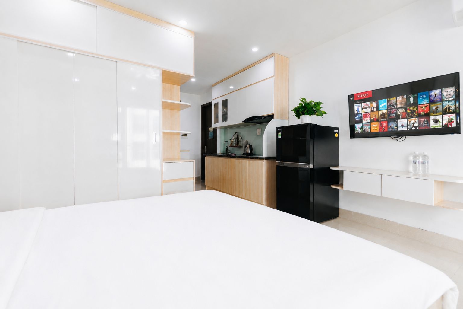 A modern bedroom with a large bed, a kitchenette with a refrigerator, and a wall-mounted TV displaying a movie selection.