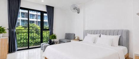 Een moderne slaapkamer met een groot bed, een grijs gestoffeerde fauteuil, een tafeltje met een lamp, en uitzicht op groen vanuit een balkon.