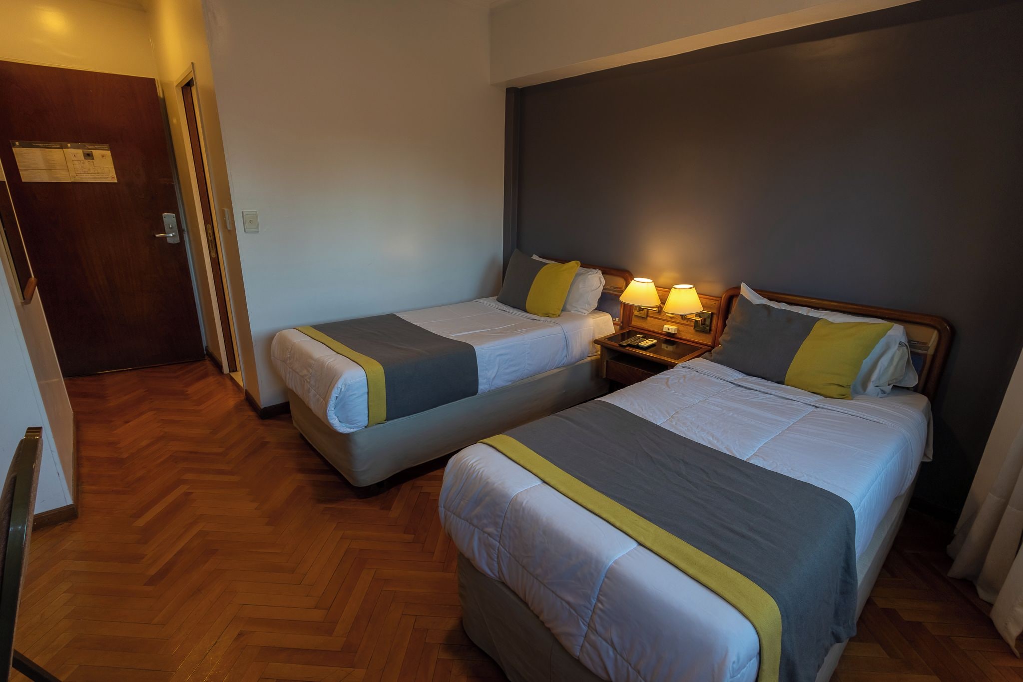Quarto de hotel com duas camas de solteiro, piso de madeira, mesa de cabeceira com luminárias e porta com placa.