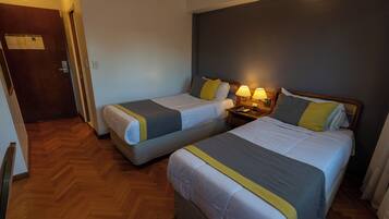 Quarto de hotel com duas camas de solteiro, piso de madeira, mesa de cabeceira com luminárias e porta com placa.