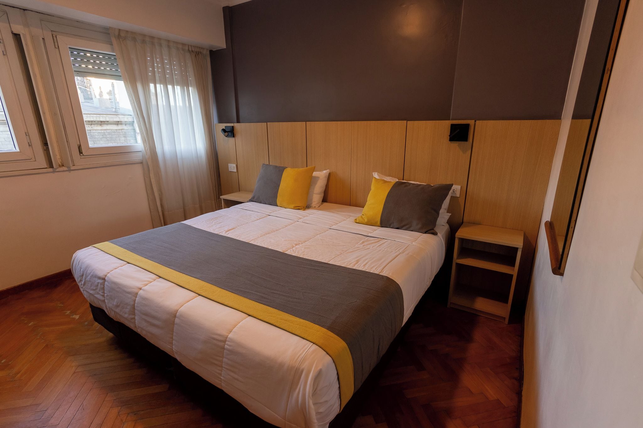 A modern hotel room with a large bed, wooden headboard, and a view of a building through the window.