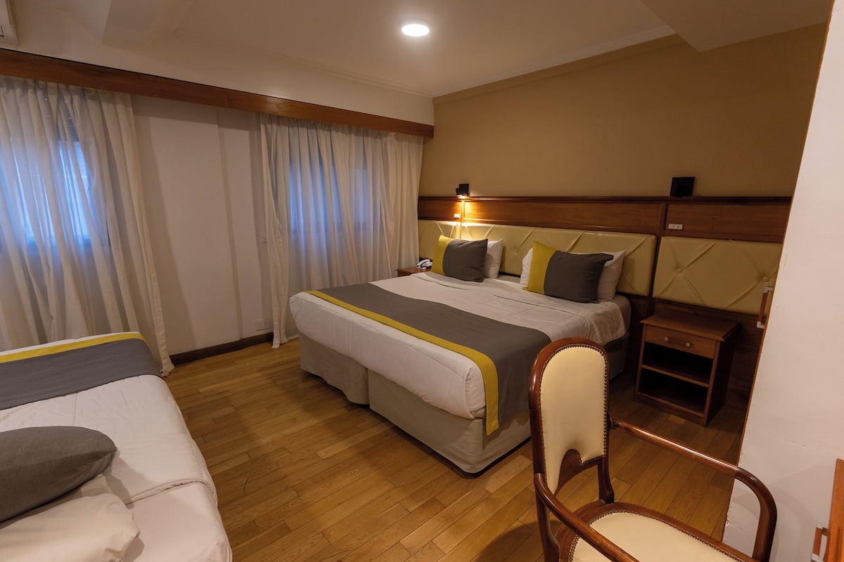 A hotel room with a large bed, a bedside table, a chair, and a window with curtains.