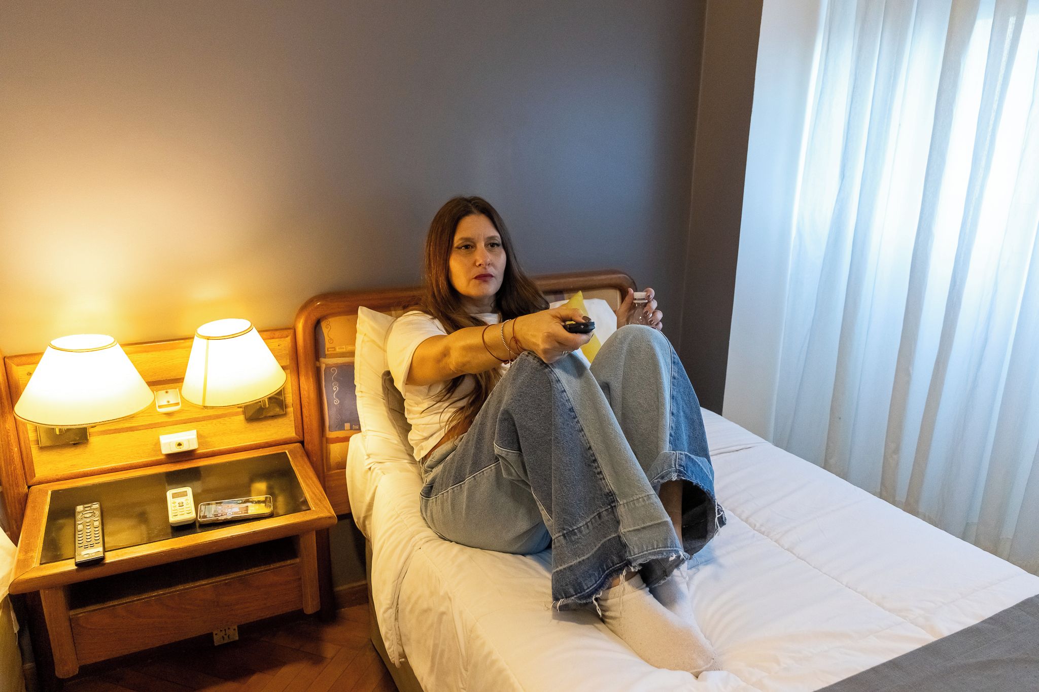 Una persona asseguda en un llit d'habitació d'hotel, amb una taula de nit, una làmpada i un comandament a distància.