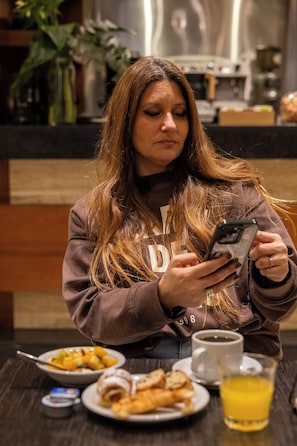Una persona sentada a una mesa con un teléfono inteligente, un plato de comida, un vaso de jugo y una taza.
