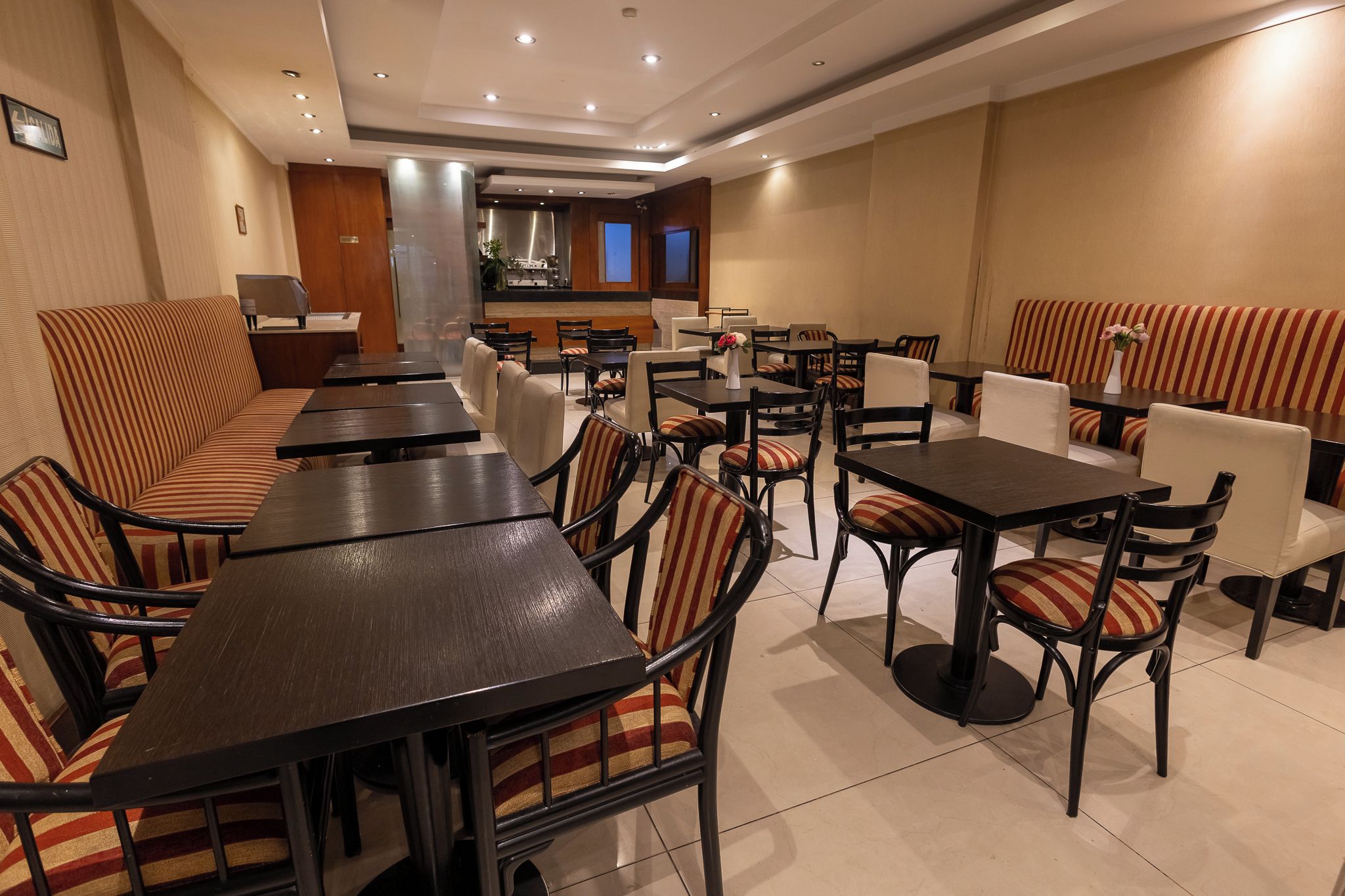 Un restaurant amb diverses taules i seients, alguns amb tapisseria estriada, i una zona de cuina al fons.