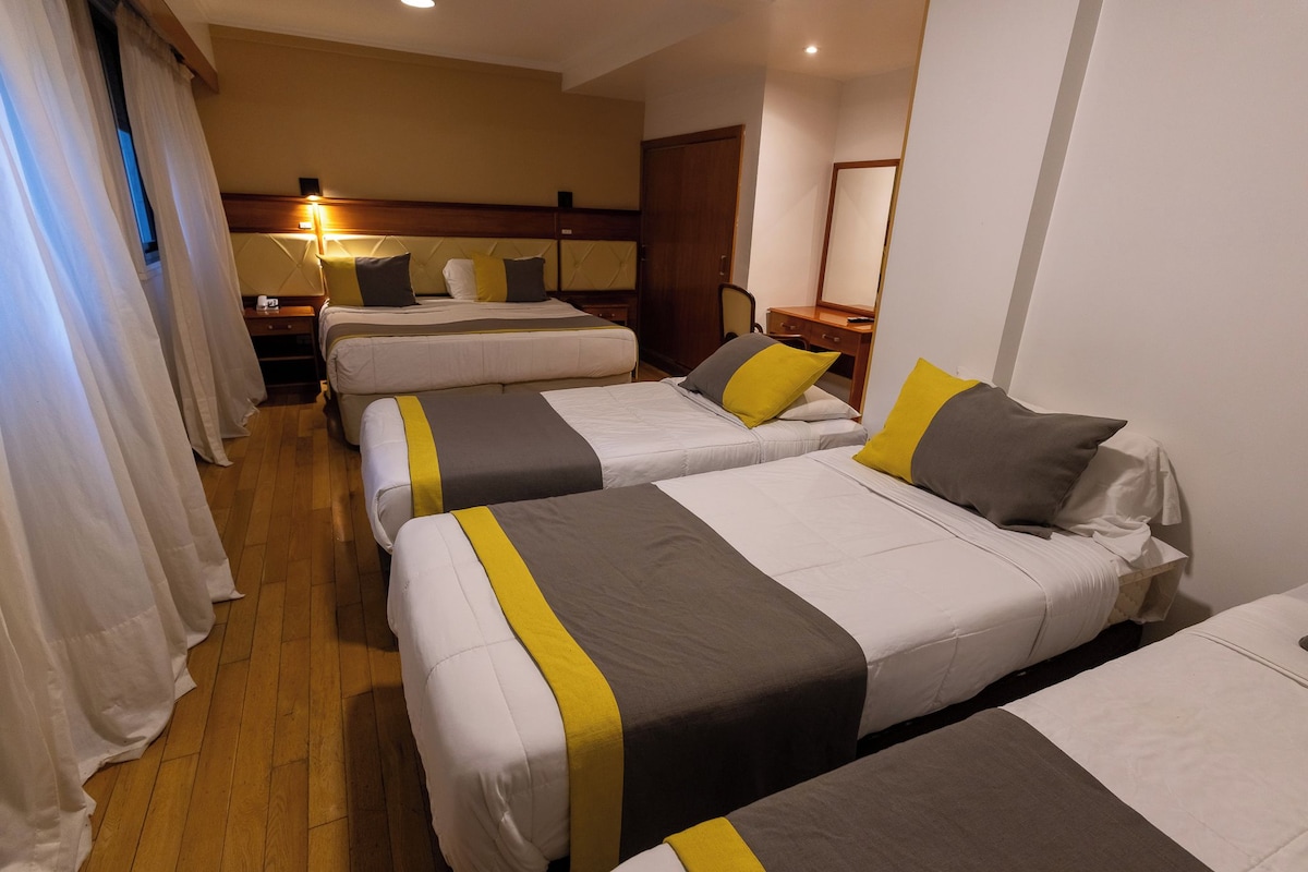 A hotel room with four beds arranged in a row, a wooden floor, a nightstand with a lamp, and a wooden door.