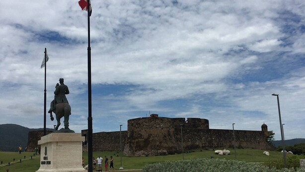 A statue on a pedestal with a flag, a stone wall, and a grassy area with flowers. - Puerto Plata | 2BR | Jacuzzi | Terrace | Modern & Central (Puerto Plata)