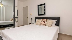 A neatly made bed with a white bedspread and decorative pillows, a wall-mounted mirror with lights, and a door in the background. - Puerto Plata | 2BR | Jacuzzi | Terrace | Modern & Central (Puerto Plata)