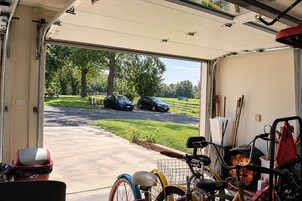 Portão de garagem aberto, com vista para a entrada da casa, dois carros estacionados e bicicletas guardadas.