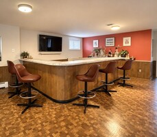 Uma área de bar com balcão curvo, quatro banquetas, uma televisão presa na parede e obras de arte emolduradas na parede vermelha.