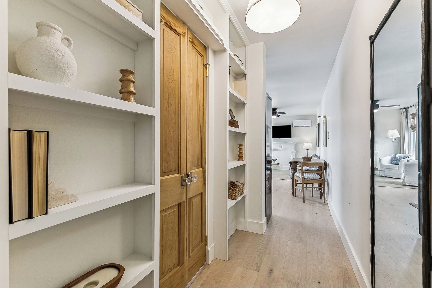 A narrow hallway with white shelving units, a wooden door, and a dining area with a table and chairs.