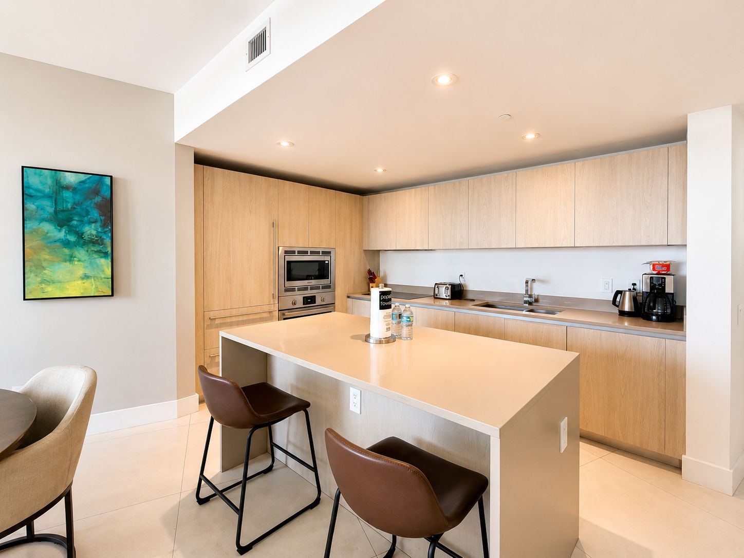 A modern kitchen with wooden cabinets, a central island with bar stools, a built-in oven, and a coffee machine.