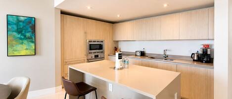 A modern kitchen with wooden cabinets, a central island with bar stools, a built-in oven, and a coffee machine.