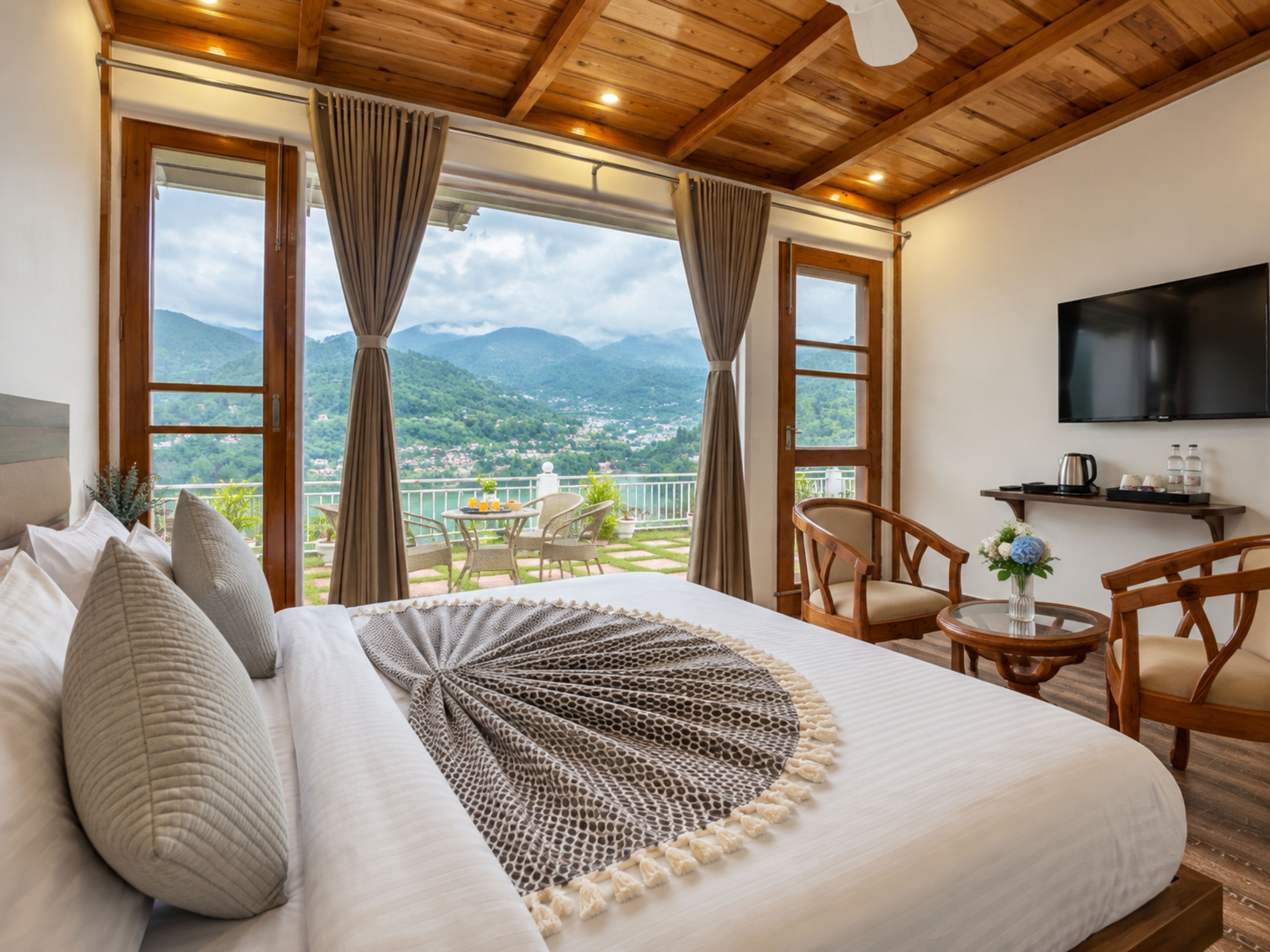 A hotel room with a bed, a TV, a balcony with a table and chairs, and a view of the mountains.