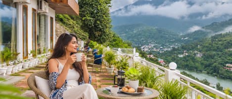 Una mujer sentada en una silla de mimbre en un balcón con vistas a un río y montañas.
