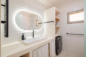 A bathroom with a white tiled wall, a large oval mirror, a white sink with a black soap dispenser, and a washing machine. - Majorelle Retreat Marrakech City Center (Marrakech)