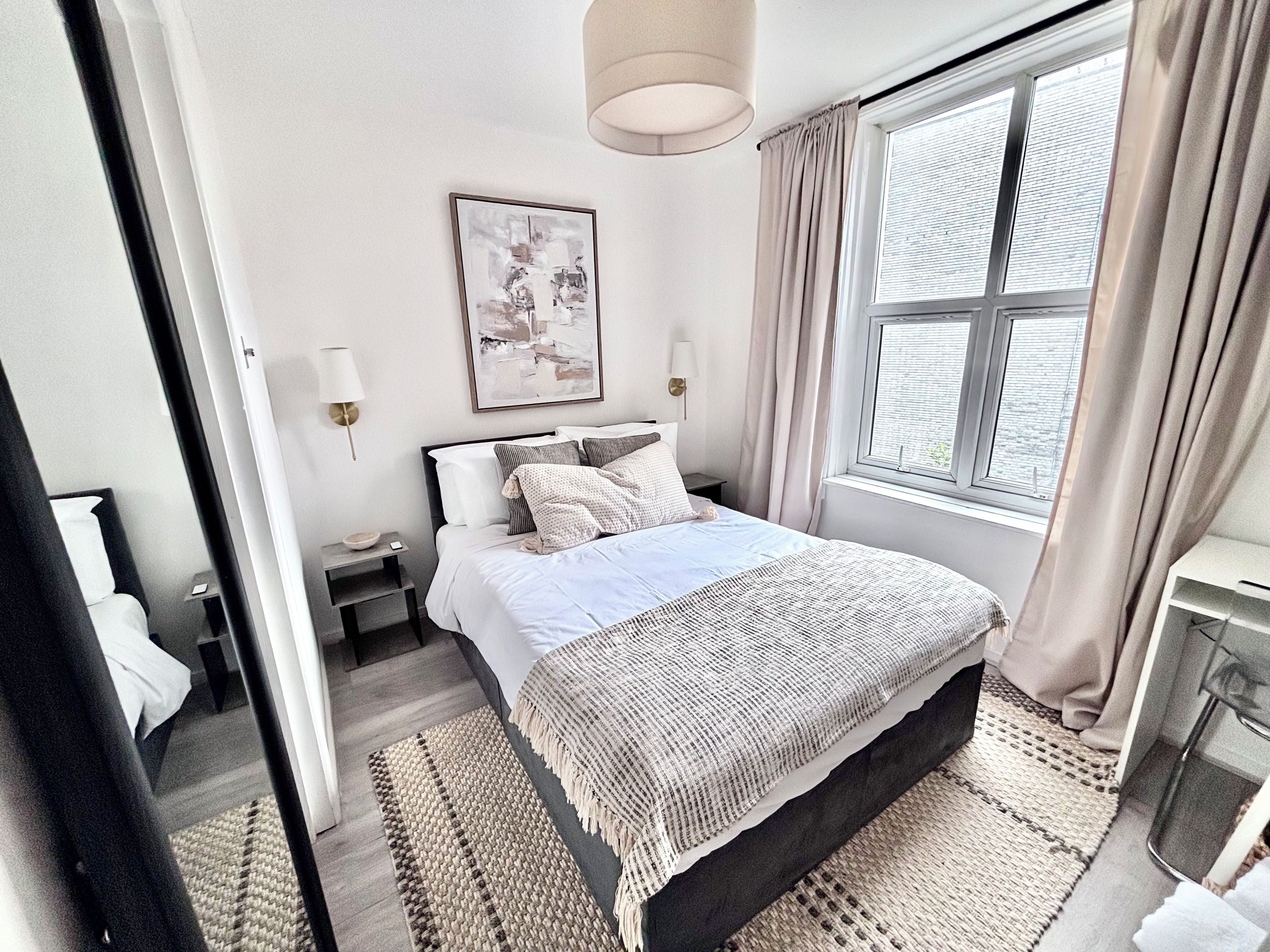 A bedroom with a bed, bedside tables, a lamp, a window with curtains, and a wall-mounted artwork.