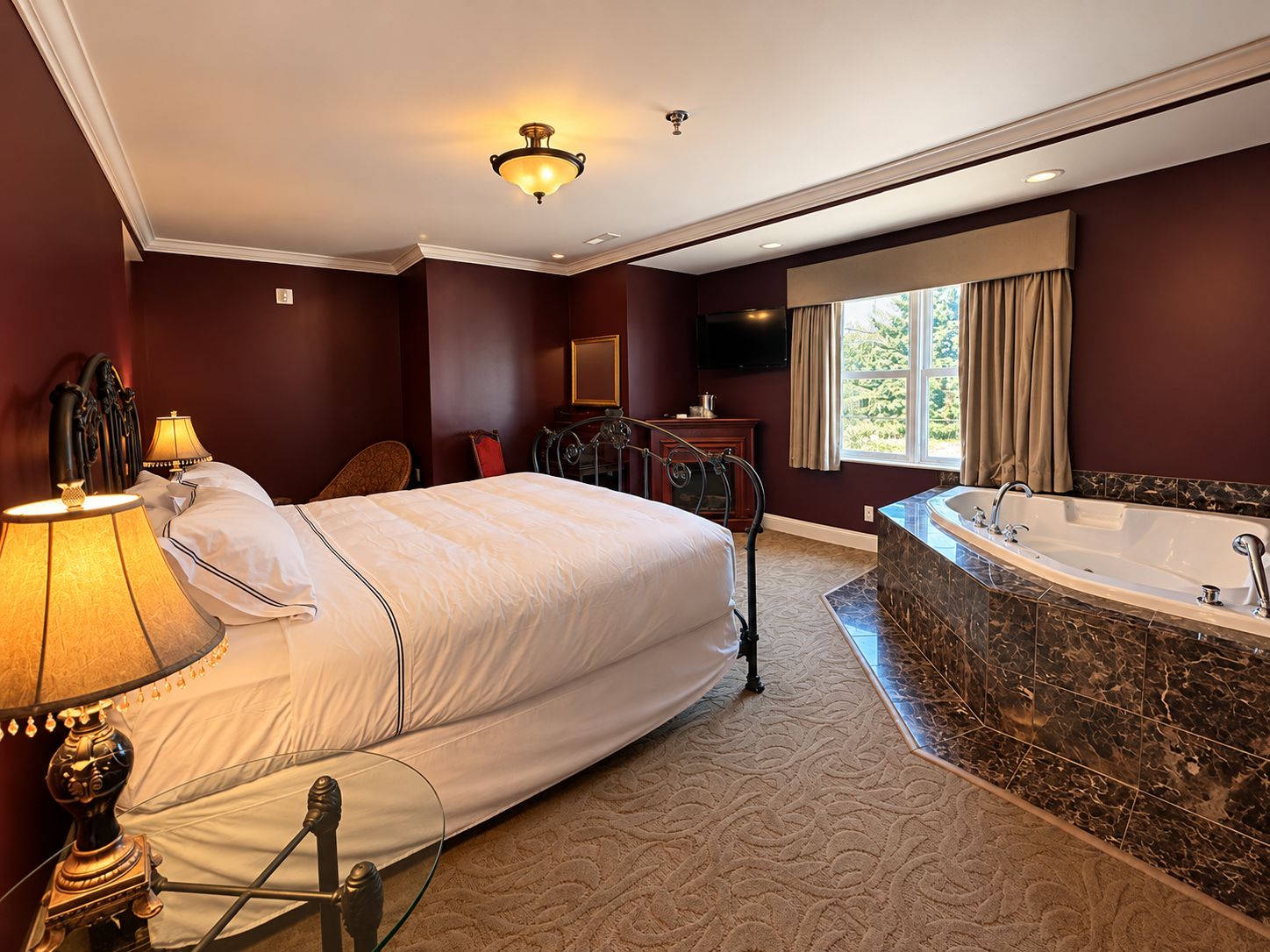 A hotel room with a large bed, a sitting area with a chair, a television, a bathroom with a bathtub, and a window with curtains.