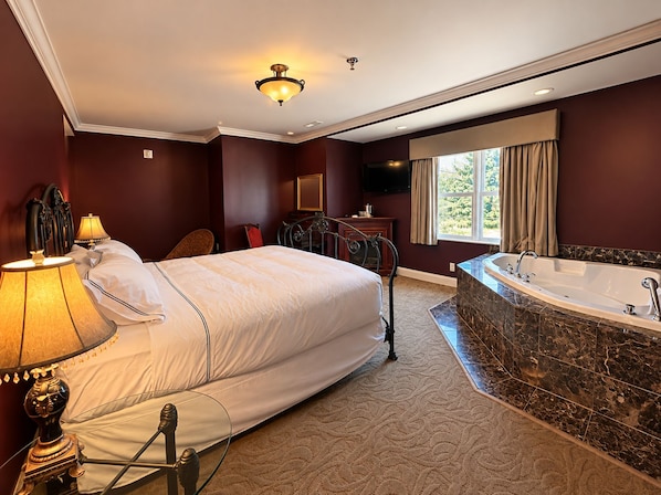 Habitación de hotel con una cama grande, una zona de estar con una silla, un televisor, un baño con bañera y una ventana con cortinas.