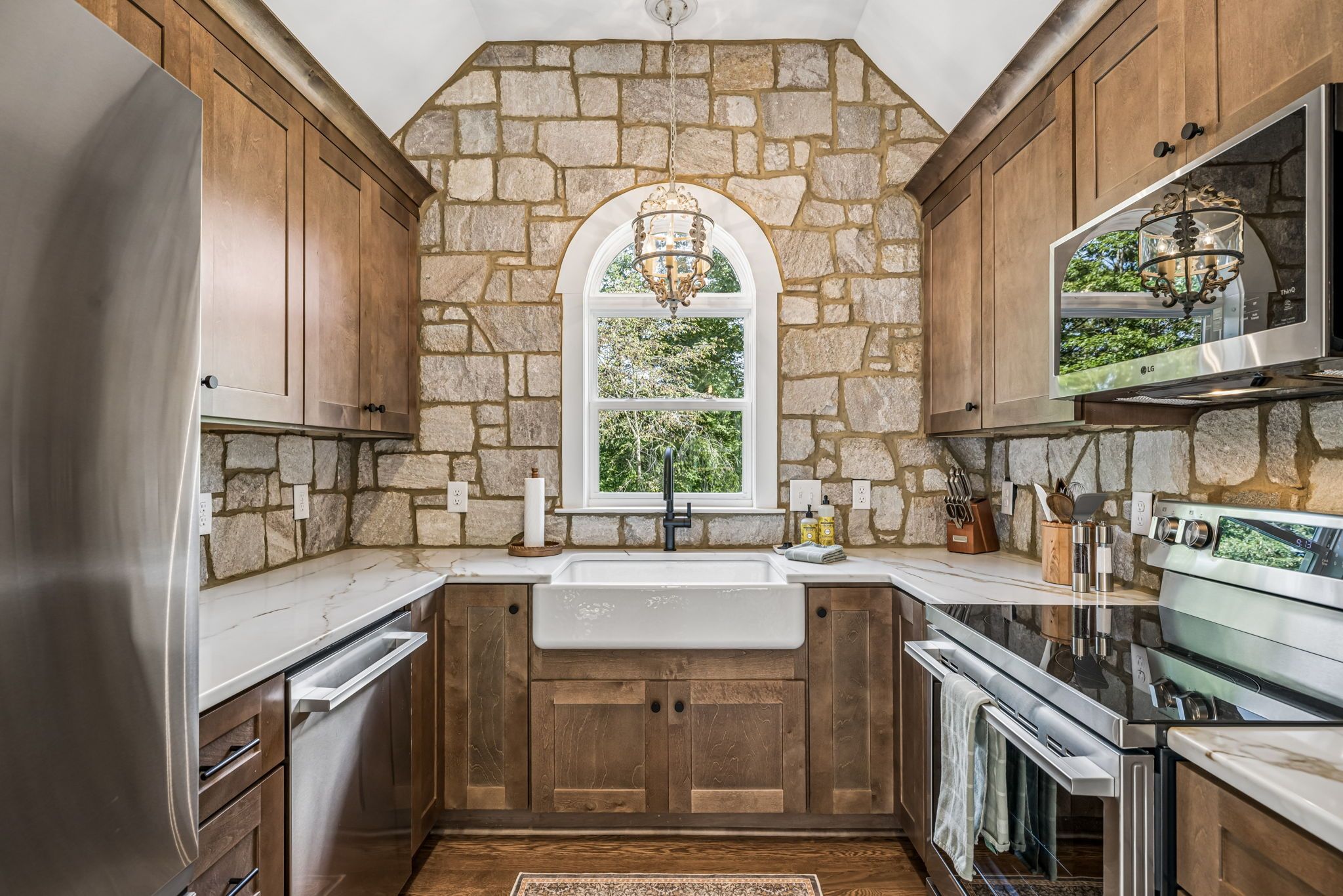 Una cocina con paredes de piedra, armarios de madera, un refrigerador de acero inoxidable, un horno y una ventana con vistas a zonas verdes.