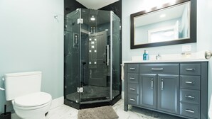 Un baño moderno con ducha envuelta en vidrio, inodoro blanco y tocador gris oscuro con varios cajones.