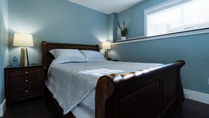 Un dormitorio con una cama de madera, dos mesitas de noche, una mesita auxiliar, una ventana con persianas y una planta en maceta en el alféizar.