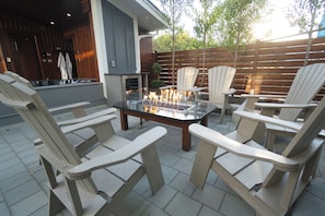 An outdoor patio area with a glass-top table surrounded by Adirondack chairs, a fire feature, and a kitchen area in the background.