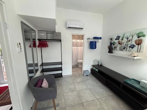 Un espace de vie compact comprenant un lit superposé, un petit coin salon, des toilettes et un climatiseur fixé au mur.