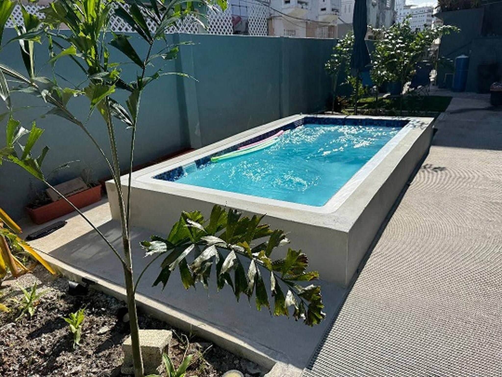 Una piscina rectangular con borde de hormigón, rodeada por un muro bajo y vegetación.