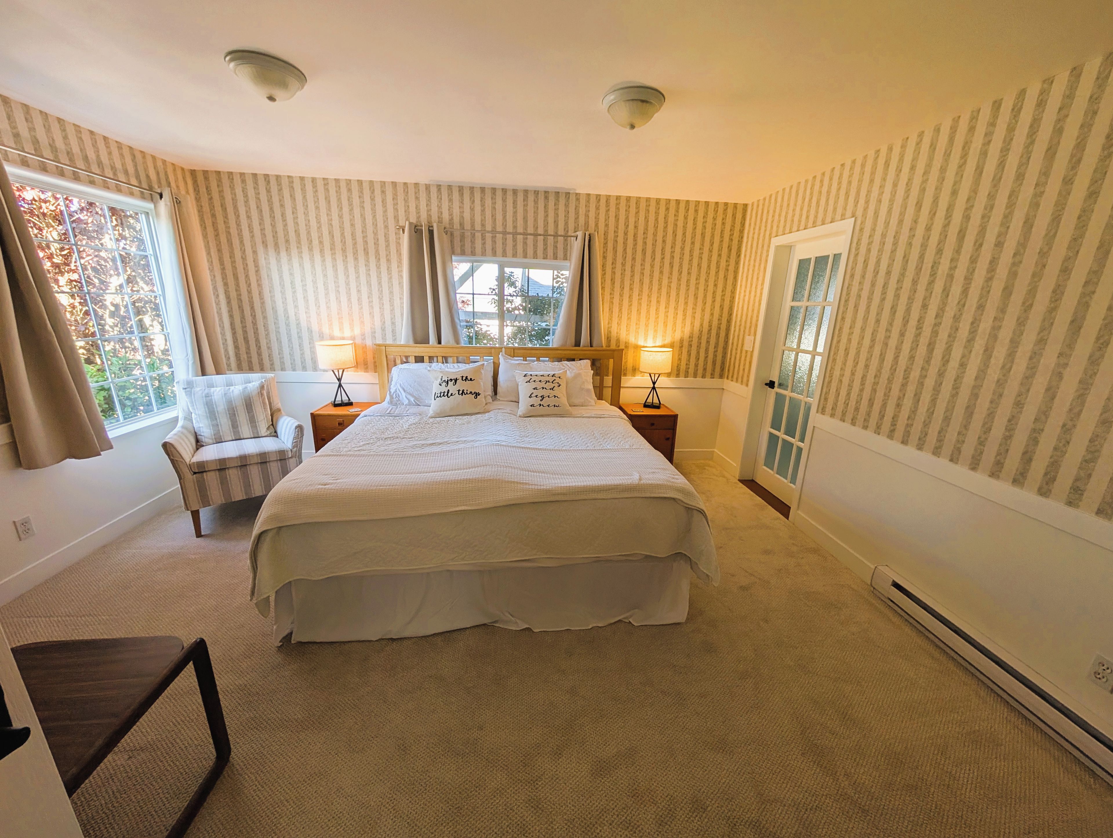 A bedroom with a large bed, two bedside tables, a chair, a window with curtains, and a door with a glass panel.