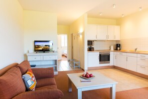 A modern living room with a sofa, a coffee table, and a television. There is a kitchen area with cabinets, a microwave, and a stove. - Ferienwohnung fur Zwei Personen, Dierhagen (Ostseebad Dierhagen)