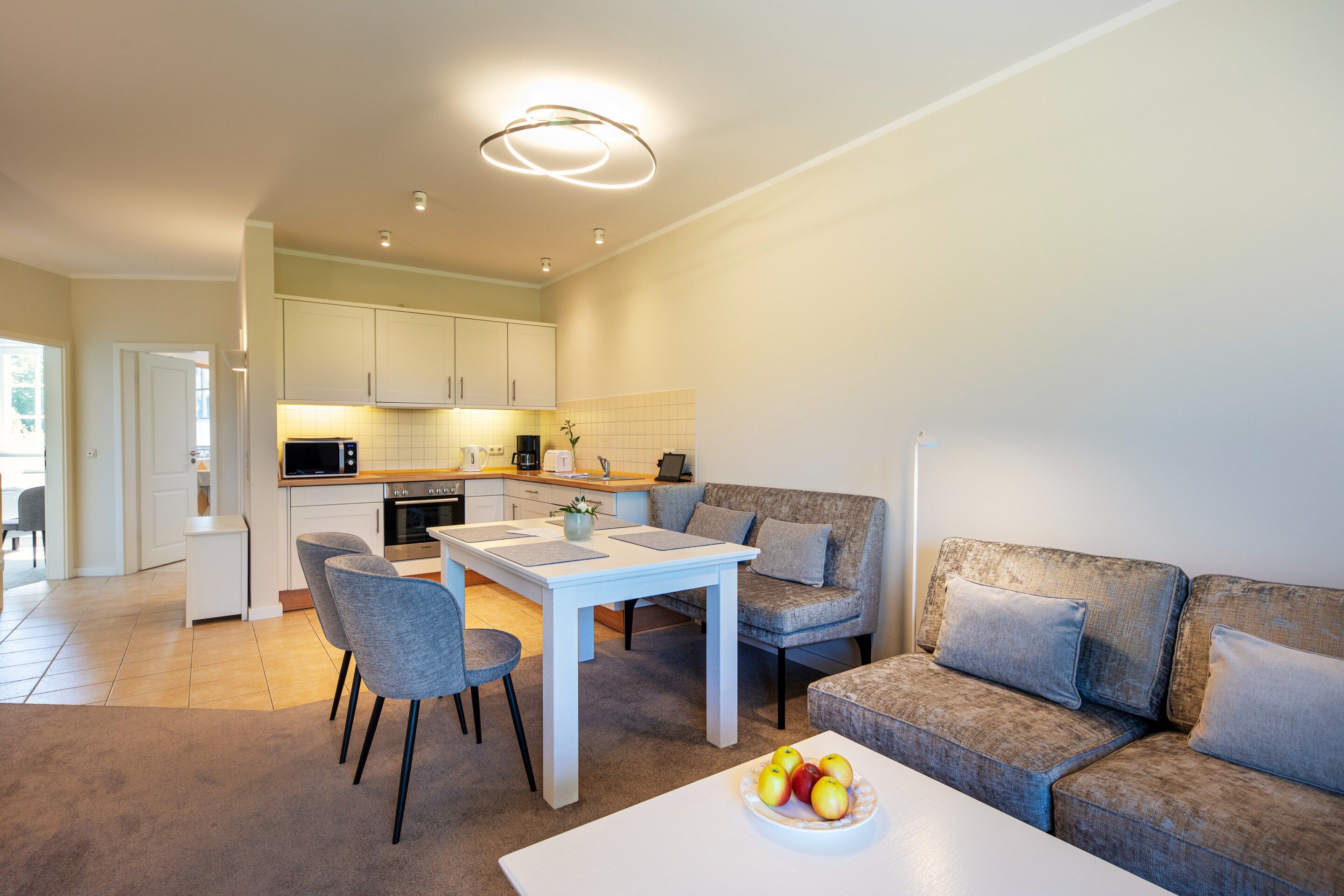 A modern kitchen with white cabinets, a dining area with a white table and chairs, and a grey sofa.