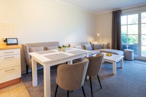 A modern living room with a sofa, a dining table with chairs, a small kitchen area with a fridge, and a large window with curtains. - Ferienwohnung fur Zwei Personen, Dierhagen (Ostseebad Dierhagen)