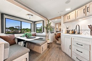 A compact RV interior with a dining area, kitchenette, and a window offering a view of the outdoors. - Glamping Getaway - On the 290 Wine Trail (Fredericksburg)