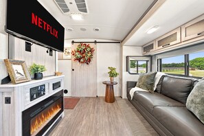 A cozy RV interior with a fireplace, a Netflix sign, a couch, and a view of the outdoors. - Glamping Getaway - On the 290 Wine Trail (Fredericksburg)