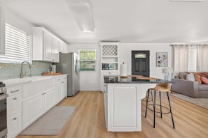 A modern kitchen with white cabinetry, a central island, and a dining area with a grey sofa and bar stools. - Lakeview Nook - 6 min to Cypress Creek Park (Austin)